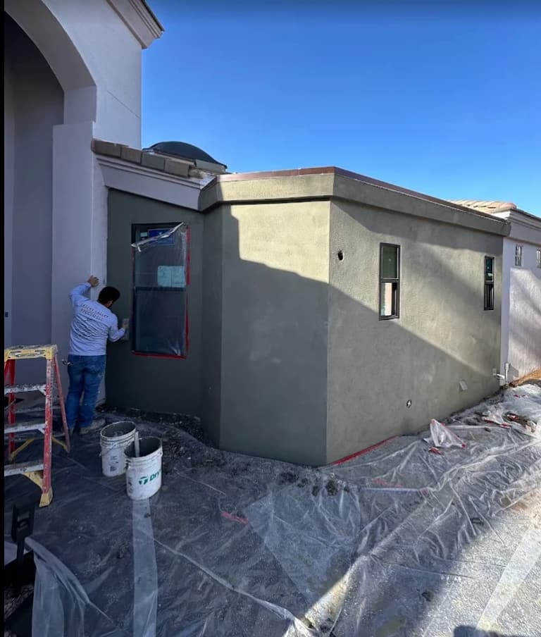 Plasterer applying an olive-green stucco finish coat to an addition, with windows masked and the ground covered in plastic