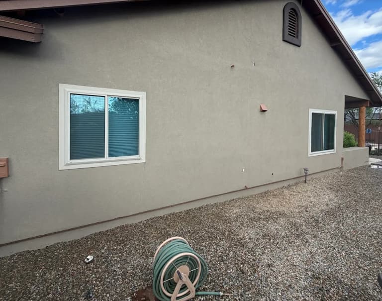 Gray stucco side wall of a single-story home with two white-framed windows and a gravel yard