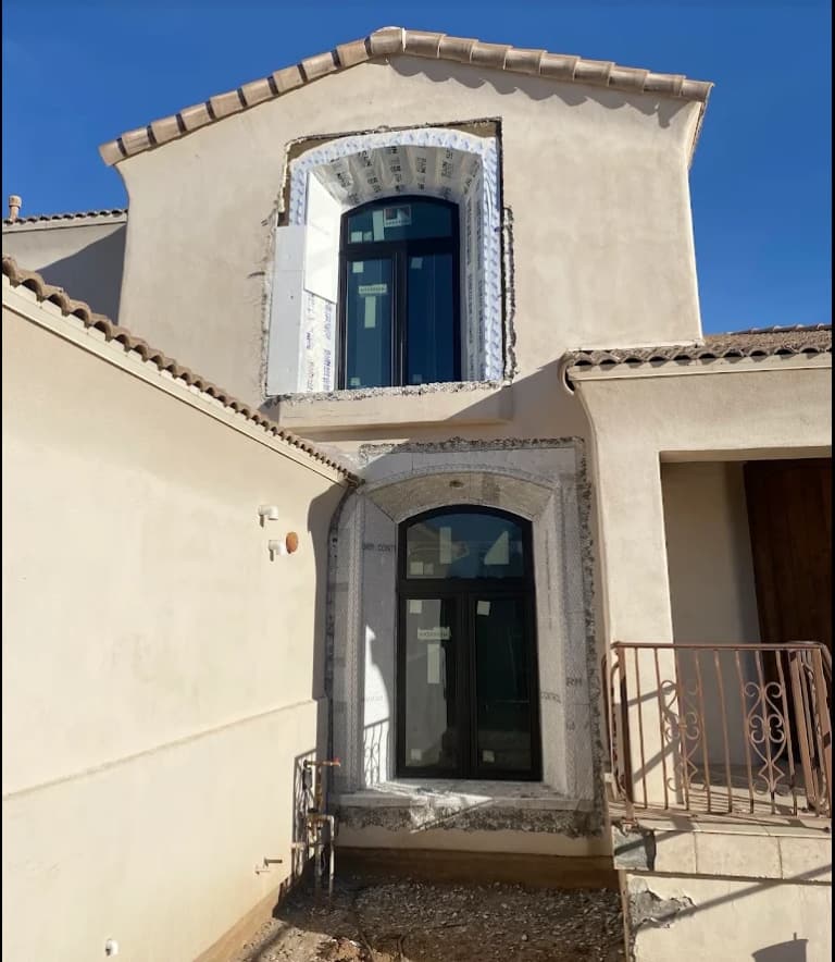 Two-story tan stucco home with stucco cut back around two new arched windows, lath exposed and ready for repair